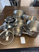 Various sized stainless steel pots with glass and metal lids displayed on a table.