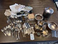 Photo shows assortment of silverware including spoons, forks, knives, various silver bowls and trays, an ice bucket with lid and wooden handle, and a pile of linens on a wooden table.