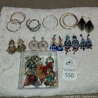 Full lot of miscellaneous earrings arranged on a soft white surface showing various hoops, dangle earrings with beadwork and filigree detail, and a clear plastic box with more earrings inside.