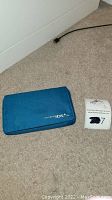 Blue Nintendo DSi XL closed with blue carrying case beside it on carpeted floor
