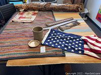 Lot items overview on table showing two rugs, American flag, woven wall art, crock, and Tapa cloth
