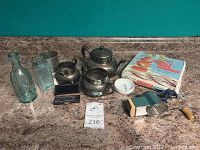 Wide view of the lot showing the silver plate tea set pieces, bottles, Kranse Kake former box, almond grinder, ashtray, stein, object micrometer, and other small items on countertop.