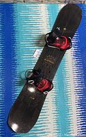 Full snowboard with attached bindings laid out on patterned blue textile background showing the entire length and top surface with bindings attached and visible straps and buckles.