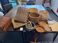 Wide view of various baskets on a table showing woven picnic basket, tall rectangular woven baskets, round baskets, flat woven trays, and small lid basket.