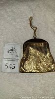 Gold colored mesh vintage bag with black top frame and wrist strap displayed on white carpet next to lot number card.