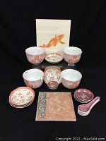 Front view of the entire dish set showing 4 large coral themed soup bowls, 5 small dishes, smaller miscellaneous round dishes, 4 ceramic spoons, and square trivet with metal decorative panel.
