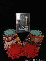 Photo showing the Kuhn Rikon stainless steel cookie press in a plastic case with multiple metal press templates, two children's baking books with colorful covers, and four intricately cut trivets in red and turquoise.