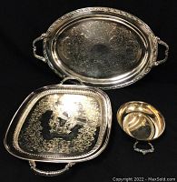 Three silver serving pieces displayed on black background: large oval tray, large square tray, small round bowl with handles. Clear view of engraved details and handle designs.