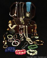 Overview of lot showing wooden jewelry box open upright with multiple necklaces draped over it and numerous bracelets arranged in front on a black surface.