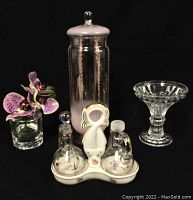 Wide view showing tall light pink gradient glass jar with lid, clear glass dish, faux orchid in glass container, and vintage mini pitchers in floral decorated ceramic holder.