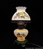 Front view of the vintage milk glass hurricane lamp with floral designs on the shade and base, showing ruffled edge and brass fittings.