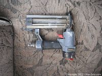 Concept 18 gauge air brad nailer with gray metal body, black textured grip, and air hose connector at the bottom, placed on patterned fabric.