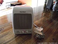 Front view of white Maxi-Heat ceramic space heater showing metal grille and control dials on wooden surface.