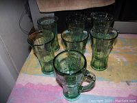 Seven green Coca-Cola glasses including one handled mug style, arranged on a towel.