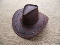 Dark brown leather cowboy hat with zigzag and cross-stitch detailing on the brim and crown, placed on a wood surface.