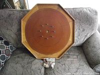 Octagonal vintage crokinole board resting on a gray couch with a bag of new wood disks placed in front. Board shows some wear and has a brown playing surface with wooden pegs near the center.