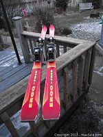 Full view of both Kneissl Ergo-Race skis with red and yellow coloring, displayed lengthwise on wooden railing outdoors on a wintery ground.