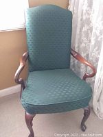 Green upholstered armchair with high back and carved wood arms and legs, positioned near a window and lace curtain.
