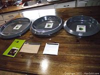 Three GreenPan pans (14", 12", 10") arranged on a wooden counter alongside product literature and packaging.