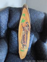 Oval live edge wood slab with oriental-style engraved figure of a person holding a staff, decorated with green leaves and grass, shown on a blue patterned sofa.