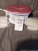 Photo of the white and pink Revlon Moisture Luxury paraffin bath unit with several packages of paraffin wax and an instruction manual displayed on a surface.