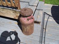 Photo of vintage metal milk/cream can sitting on painted wooden decking with shadows cast around it, showing rust and patina on the surface and the engraved text on the side.