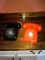 Side by side image of a black and orange rotary dial telephones on a wooden surface.