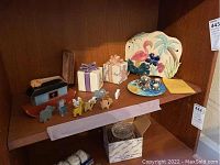 Shelf display showing three music boxes and wooden Noah's Ark animal figures along with small ceramic and glass figurines and flamingo-themed plate.