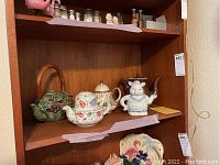 Five tea pots and one matching cup on wooden shelf, seen with floral and character designs