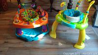 Two baby bouncers on wooden floor; orange and blue one with colorful toys, green and yellow one with cushioned blue seat and toys.