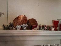 Shelf display showing woven fan decorations, a glass vase in red, a white ceramic vase, small Christmas figurines, and additional candle holders and décor items arranged linearly.