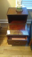 Full view of vintage wooden end table showing glass-covered leather inlay top with gold decorative edge, open shelf, single drawer with brass handle pulled out to display interior, standing on wooden floor.