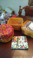 Three Longaberger baskets on table including two uncovered woven baskets and one covered with a colorful floral fabric liner and lid, plus folded floral cloth nearby.