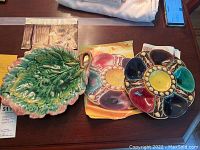Two pieces of maiolica pottery displayed on a wooden table including a green leaf-shaped serving tray with a handle and a colorful segmented oyster bowl with yellow center.