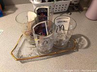 Four clear glass McDonald's Disney World 25th anniversary limited edition glasses, each with unique designs and official rules text, all placed on a hexagonal glass serving platter with gold trim on a countertop.