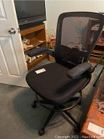 Front-left angle view of the black office chair showing padded upholstered seat, mesh backrest, and adjustable padded armrests. The chair is on a carpeted floor near a wooden chest and an entertainment center.
