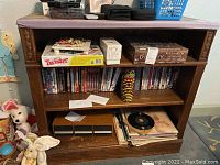 Wooden shelving unit containing records, cassette tapes, DVDs, and boxed games including Twister and Duck Dynasty Redneck game, with miscellaneous items such as a striped cup and toys nearby.