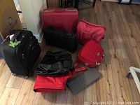 Displays all eight pieces of luggage including the red hard shell suitcase, black and red rolling suitcases, various soft bags in red and black, and a gray case on a wooden floor.