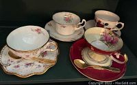 Four teacup sets with matching saucers and spoons, displayed side by side on a shelf, showing floral and gold trim patterns, one saucer visibly chipped.