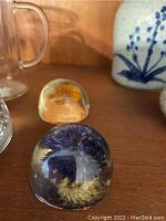 Close-up of two hand blown glass paperweights; one has orange and yellow flower inside, the other purple flower inside.