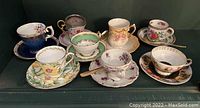 Photo showing eight assorted vintage tea cups and saucers with floral and gold trim designs arranged on a dark surface.