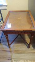 Front view of vintage wooden table with glass top and wood inlay showing central floral design and carved legs.