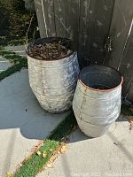 Photo showing two large weathered metal garden pots with ribbed barrel-like design, one containing dried leaves, placed outdoors near a building with concrete and grass around.