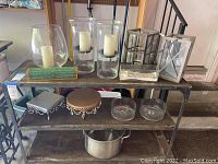 Three shelves with glass candle holders containing pillar candles, decorative platters, small glass bowls and a large metal pot below.