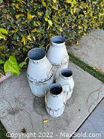 Four white distressed metal pots in varying sizes placed on a concrete surface next to greenery.