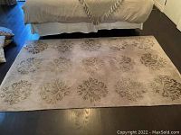 View of full 5x7 area rug laid on dark hardwood floor under a bed, beige color with brown leaf motifs spaced evenly