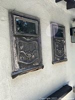 Pair of weathered, distressed antique-style wall mirrors mounted on a light exterior wall. The frames have carved detailing and paint chipping, with a small shelf at the base of each mirror.