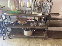 Full view of the metal and wood bar cart showing three shelves, metal frame with rivets, caster wheels, and various items placed on the bar cart for scale and detail.