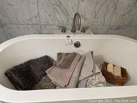 Photo showing several folded bath towels in neutral tones placed inside a bathtub along with a small woven basket holding rolled towels.