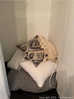 Pillows stacked in a corner including a patterned decorative pillow, cream textured pillow, grey pillow, and beige throw blanket with ruffled edge.